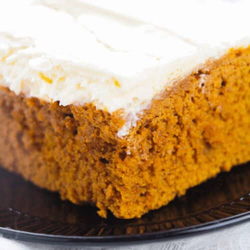 Close-up of a slice of moist pumpkin cake with a thick layer of cream cheese frosting on a black plate, evoking the comforting aromas reminiscent of pumpkin spice cake.