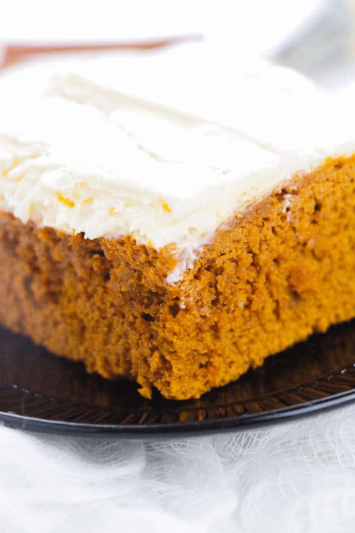 Close-up of a slice of moist pumpkin cake with a thick layer of cream cheese frosting on a black plate, evoking the comforting aromas reminiscent of pumpkin spice cake.