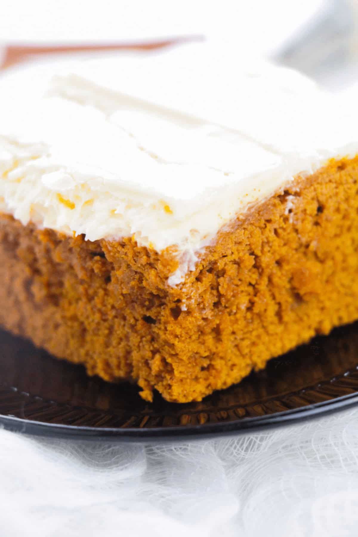 Close-up of a slice of moist pumpkin cake with a thick layer of cream cheese frosting on a black plate, evoking the comforting aromas reminiscent of pumpkin spice cake.