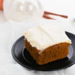 A slice of frosted pumpkin spice cake on a black plate with white pumpkin decor and cinnamon sticks in the background.