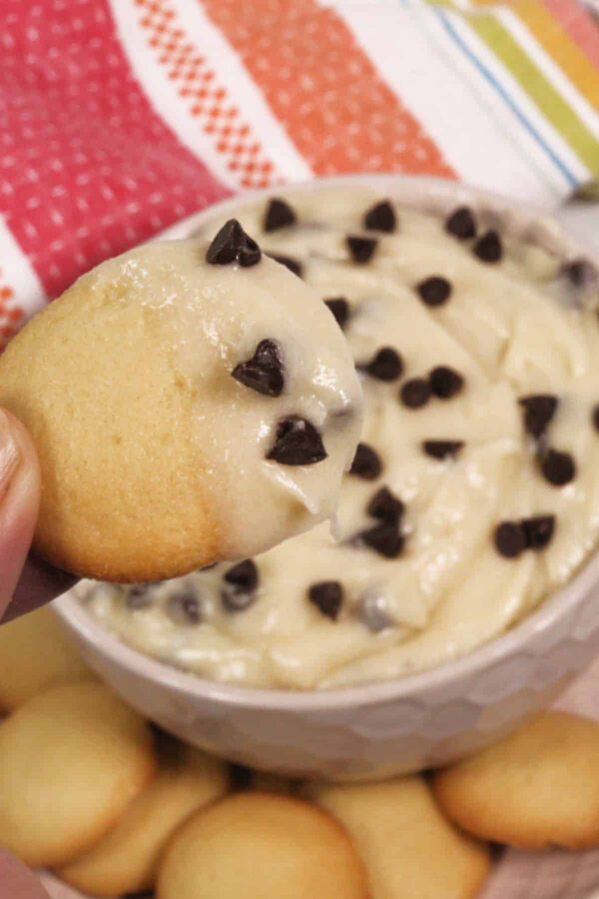 A hand holds a vanilla wafer dipped in rich Chocolate Chip Dip, topped with chocolate chips, with more wafers and a bowl of the delicious dip in the background.