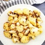 A white plate of air fryer fried cauliflower florets sits on a gray surface, with a striped towel and a bulb of garlic in the background.