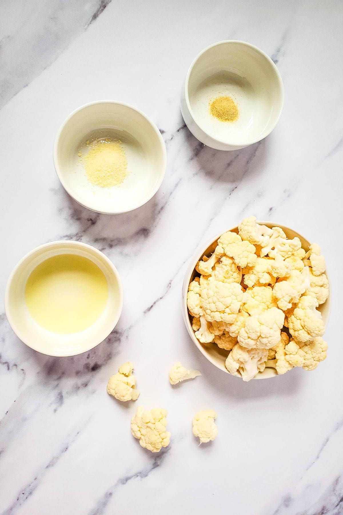 Four bowls on a marble surface hold cauliflower florets, oil, grated cheese, onion powder, and garlic powder—ingredients perfect for making crispy air fryer fried cauliflower. A few scattered florets add to the inviting prep scene.