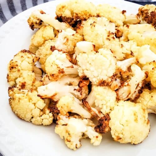 Air fryer fried cauliflower florets served on a white plate, with a striped cloth and a garlic bulb in the background.