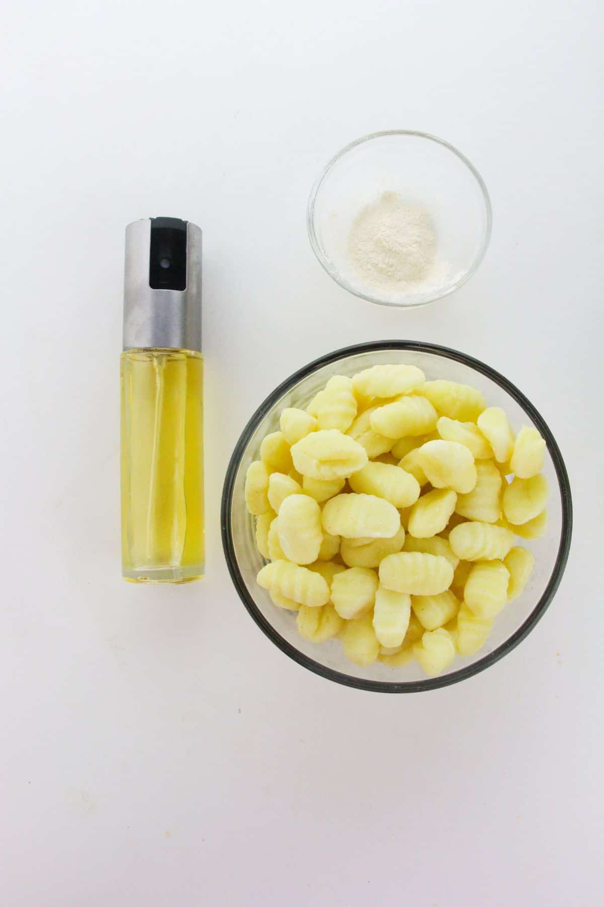A small glass bowl of uncooked gnocchi, a small glass bowl of white powder, and a spray bottle of oil are arranged on a white surface—perfect for making crispy Air Fryer Gnocchi.