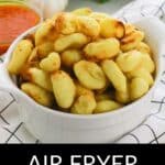 A white bowl filled with golden-brown Air Fryer Gnocchi sits on a checkered cloth, surrounded by parsley, a garlic bulb, oil, and red sauce in the background.
