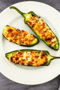 Three jalapeño peppers, transformed into delectable air fryer jalapeno poppers, are stuffed with a rich cheese mixture and perfectly browned on top, served elegantly on a white plate.