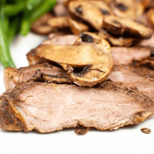 Sliced air fryer ribeye steak topped with sautéed mushrooms, served alongside green beans on a pristine white plate.