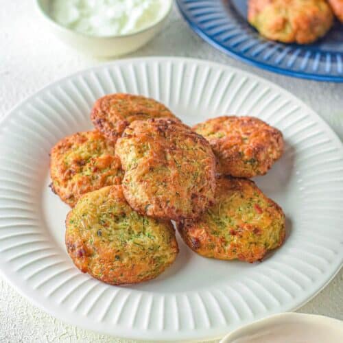 A white plate with several round Air Fryer Zucchini Fritters, accompanied by a bowl of white sauce and a bowl of red sauce on the side. More fritters are on a blue plate in the background.