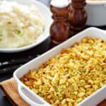 A white baking dish filled with golden Apple and Sausage Dressing sits on a mat, with a bowl of mashed potatoes, gravy, and salt and pepper shakers in the background.