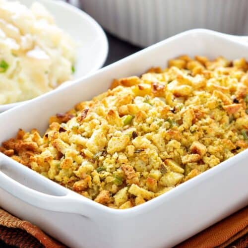 A white baking dish filled with Apple and Sausage Dressing sits on a folded napkin, with mashed potatoes and another casserole visible in the background.