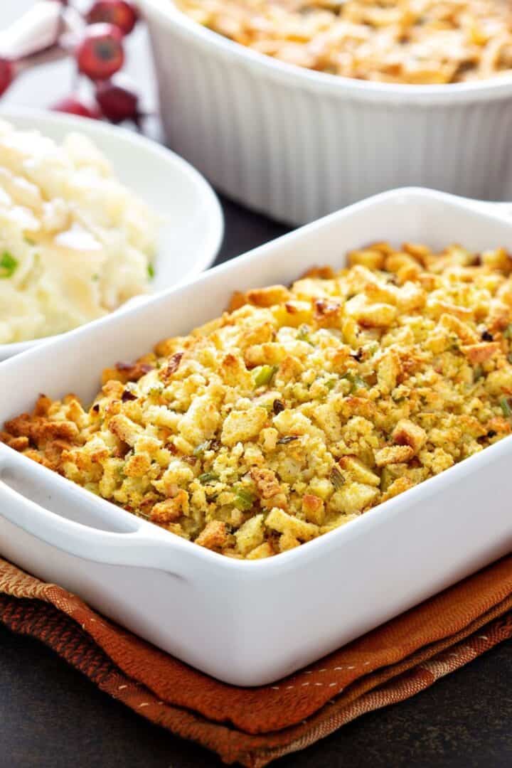 A white baking dish filled with Apple and Sausage Dressing sits on a folded napkin, with mashed potatoes and another casserole visible in the background.