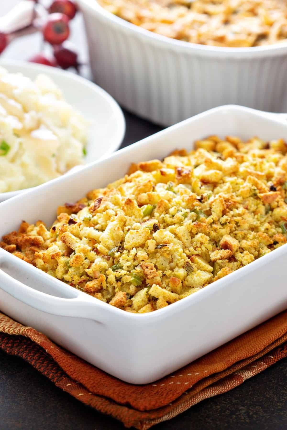A white baking dish filled with Apple and Sausage Dressing sits on a folded napkin, with mashed potatoes and another casserole visible in the background.