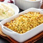 A white casserole dish filled with golden-brown apple and sausage dressing sits on a brown cloth, with a bowl of mashed potatoes visible in the background.