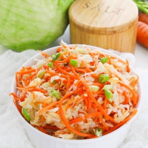 A white bowl filled with shredded cabbage and carrots sits on a white cloth, creating a fresh Carrot Slaw, with a cabbage, salt container, pepper mill, and carrot in the background.