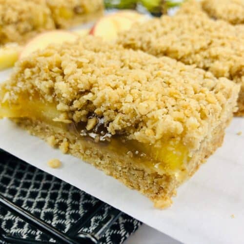 A close-up of a crumbly, layered apple oatmeal bar on parchment paper, with fresh apple slices and another apple oatmeal bar in the background.