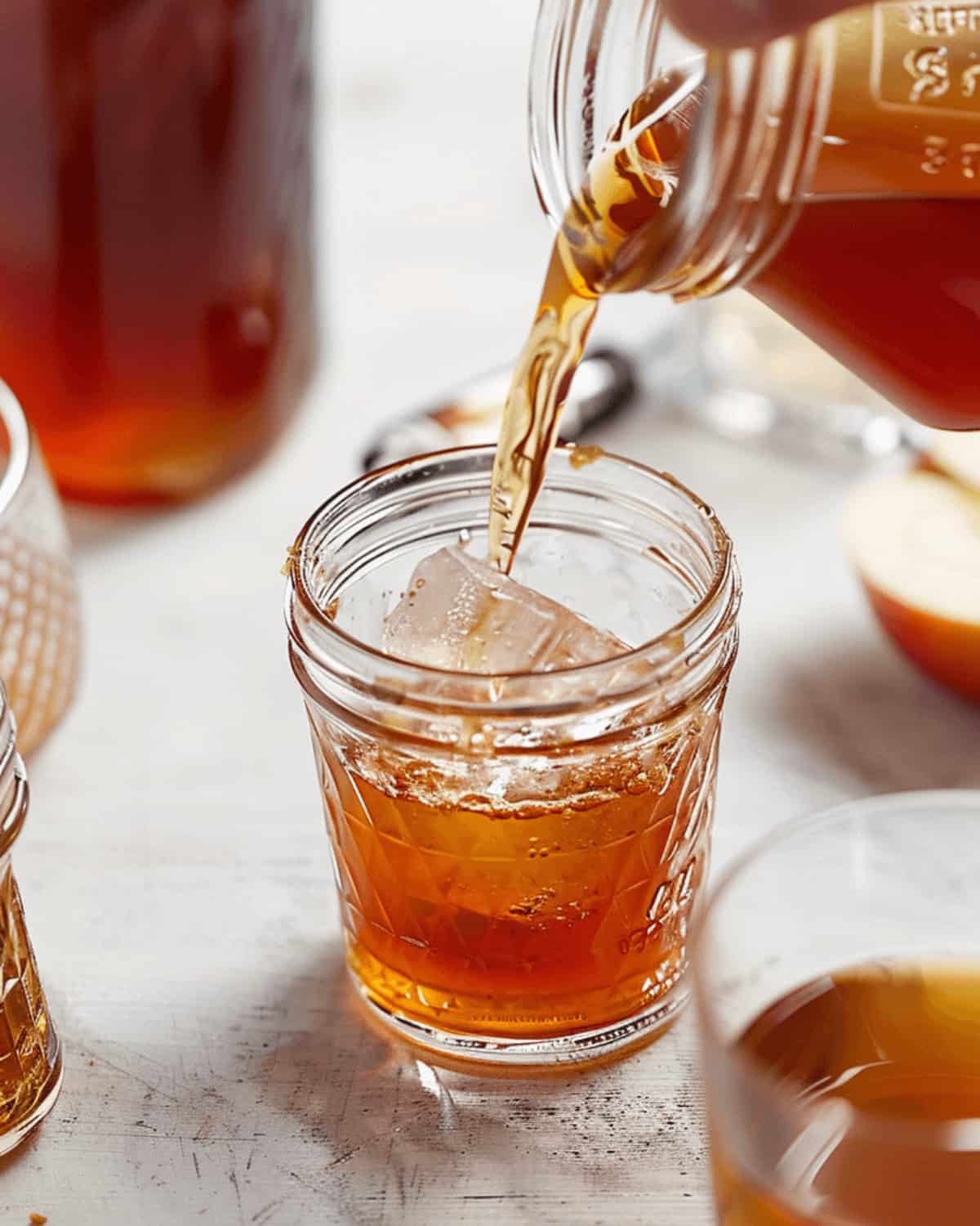 A hand pours apple pie moonshine from a glass jar into a glass filled with ice cubes on a white surface, with additional jars and a sliced apple in the background, evoking the cozy feeling of enjoying apple pie moonshine.