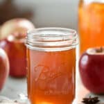 A glass jar filled with apple pie moonshine, surrounded by fresh apples and spices, sits on a countertop.