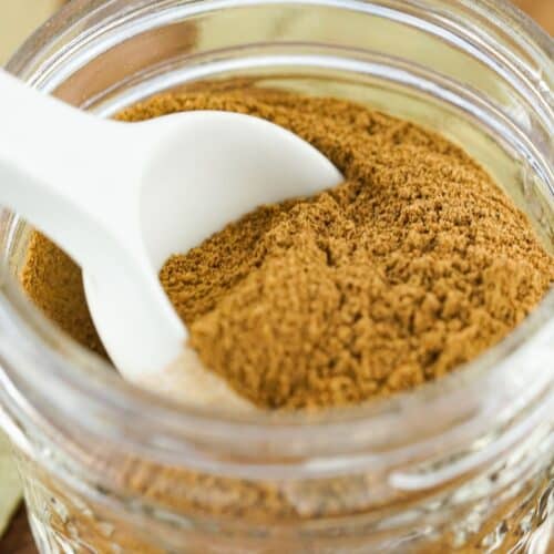 A glass jar filled with ground cinnamon and Apple Pie Spice, with a white spoon resting inside.