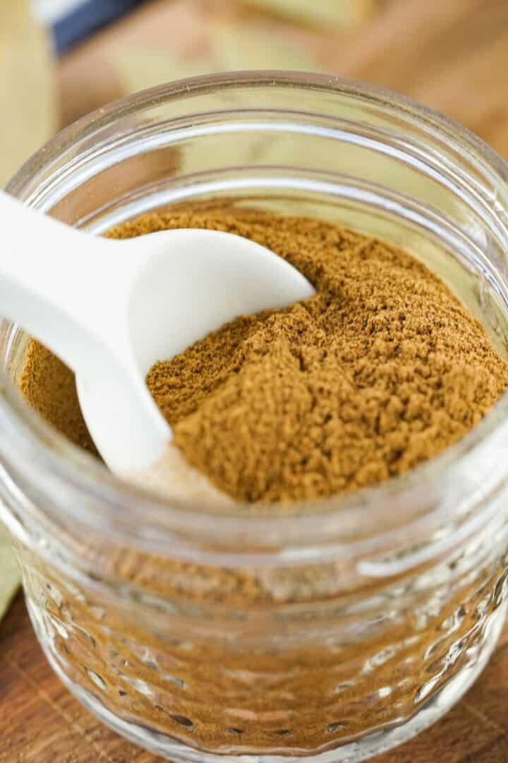 A glass jar filled with ground cinnamon and Apple Pie Spice, with a white spoon resting inside.