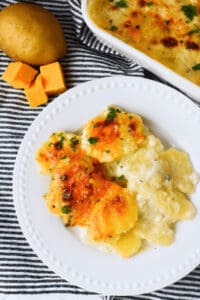 Plate of the side dish, garnished with herbs, next to cubes of cheddar and a whole potato, on a striped cloth.