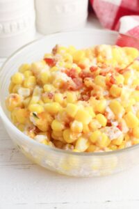 A clear bowl filled with a delightful corn casserole with cream cheese, crowned with bacon bits. The background features blurred white jars and a red checkered cloth.