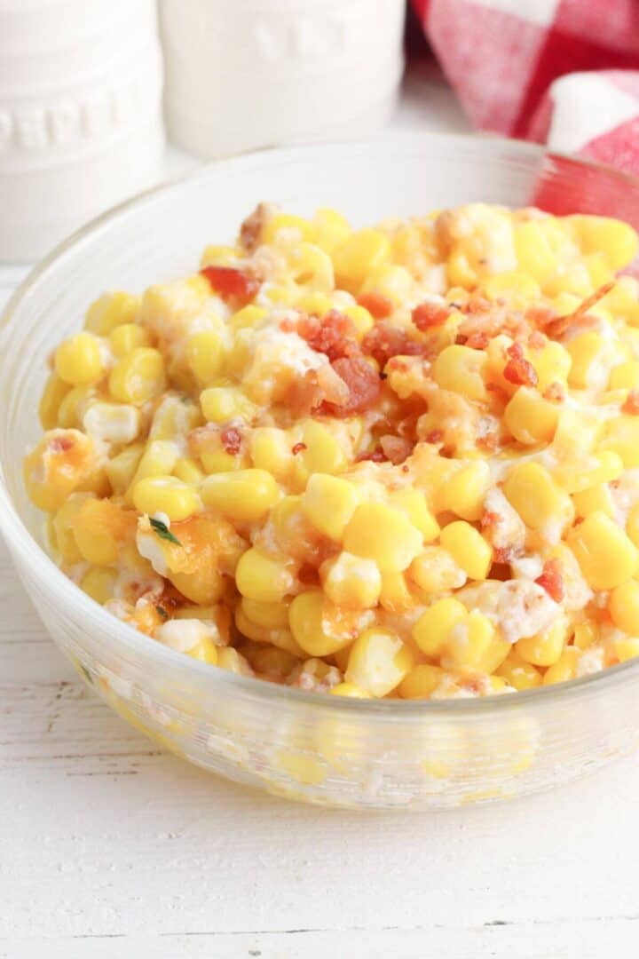 A clear bowl filled with a delightful corn casserole with cream cheese, crowned with bacon bits. The background features blurred white jars and a red checkered cloth.