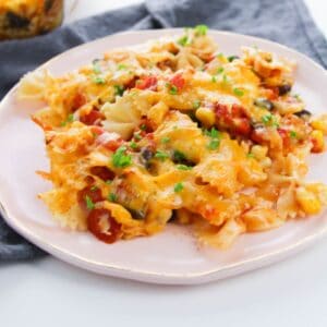 A plate of Enchilada Pasta with bowtie noodles, melted cheese, diced vegetables, and herbs, served on a light pink plate with a gray napkin underneath.