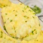 A close-up of a spoonful of Baked Twice Mashed Potatoes topped with chopped chives, with more mashed potatoes and chives visible in the background.