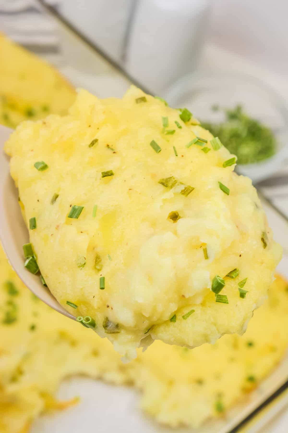 A close-up of a spoonful of Baked Twice Mashed Potatoes topped with chopped chives, with more mashed potatoes and chives visible in the background.
