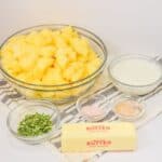 A bowl of cubed veggies, stick of butter, bowls of chopped chives, milk, and seasonings are arranged on a striped cloth.