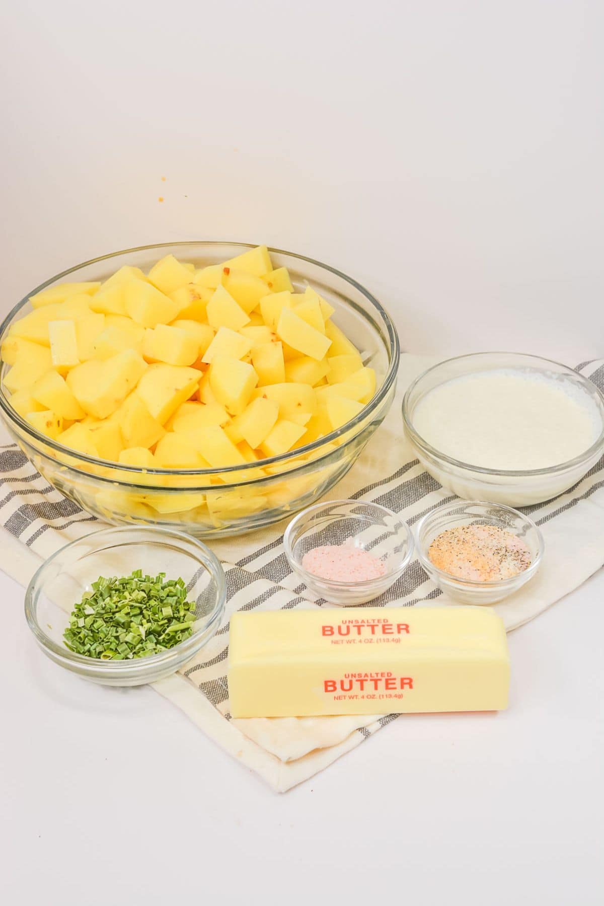 A bowl of cubed veggies, stick of butter, bowls of chopped chives, milk, and seasonings are arranged on a striped cloth.