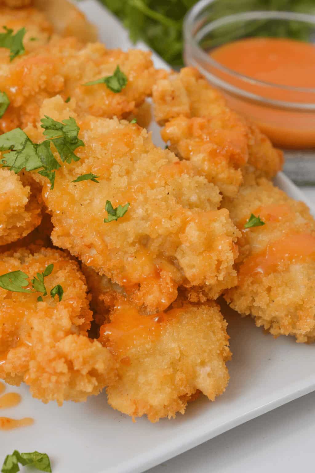 Close-up of crispy breaded Bang Bang Chicken drizzled with sauce and garnished with fresh herbs on a white plate. A small bowl of dipping sauce waits eagerly in the background.