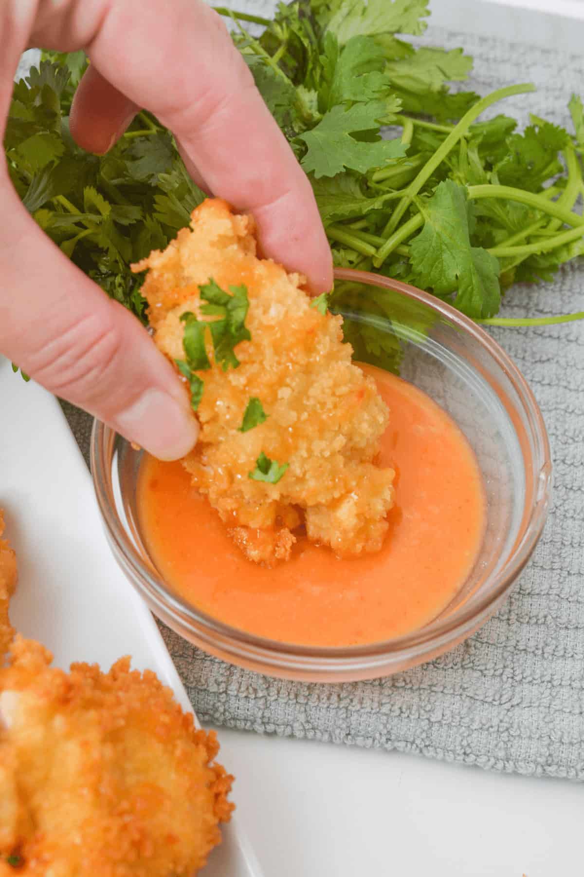 A hand dips a piece of crispy, golden fried shrimp into a small glass bowl of orange sauce resembling the spicy tanginess of Bang Bang Chicken, with fresh cilantro in the background.