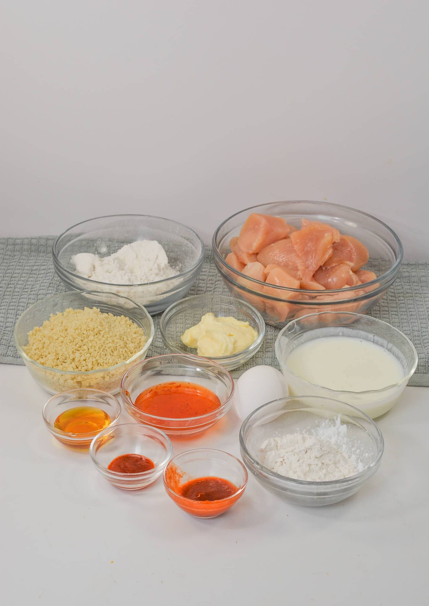 Bowls of raw chicken pieces, flour, breadcrumbs, milk, egg, butter, and various spices for preparing a delicious meal arranged on a light surface.