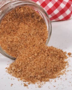 A jar spills a mix of ground cinnamon and sugar onto a white surface, with a red and white checkered cloth in the background, evoking the cozy charm of homemade BBQ rub prep.