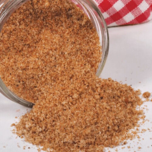 A jar spills a mix of ground cinnamon and sugar onto a white surface, with a red and white checkered cloth in the background, evoking the cozy charm of homemade BBQ rub prep.