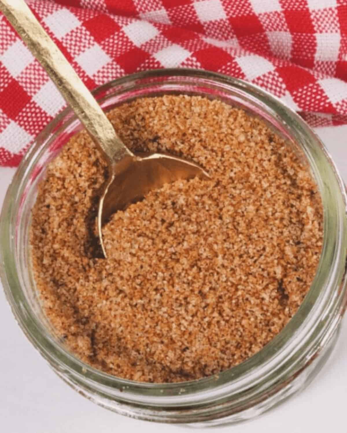 An open glass jar filled with a rich brown BBQ rub, with a gold spoon inside. A red and white checkered cloth is in the background.