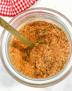A jar filled with a blend of various ground spices labeled as burger seasoning. A metal spoon is inserted into the mix, and a red and white checkered cloth is partially visible in the background.