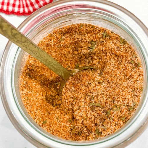 A jar filled with a blend of various ground spices labeled as burger seasoning. A metal spoon is inserted into the mix, and a red and white checkered cloth is partially visible in the background.