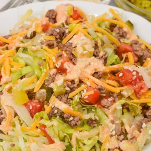 A plate of Big Mac Salad with ground beef, shredded cheddar cheese, lettuce, cherry tomatoes, and creamy dressing, accompanied by small bowls of pickles and sesame seeds in the background.