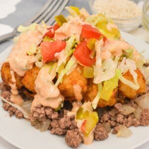 A plate of seasoned ground beef and onions, inspired by Big Mac Tater Tot Casserole, topped with tater tots, shredded lettuce, diced tomatoes, sliced peppers, and creamy sauce.