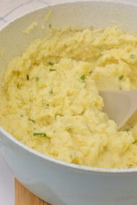 A pot of creamy Boursin Mashed Potatoes with chopped green herbs is being stirred with a spoon.