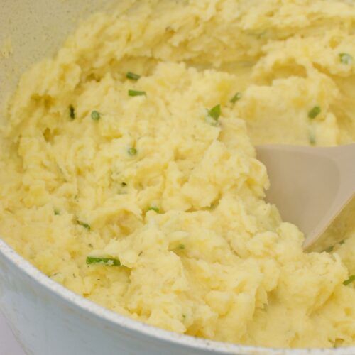 A pot of creamy Boursin Mashed Potatoes with chopped green herbs is being stirred with a spoon.