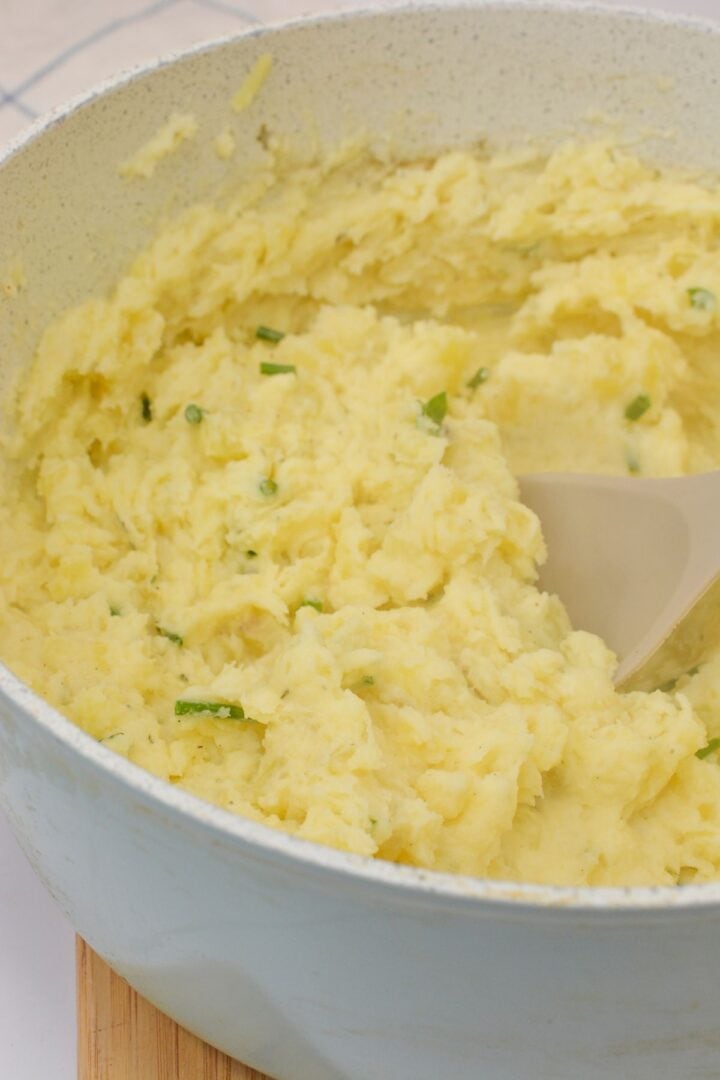 A pot of creamy Boursin Mashed Potatoes with chopped green herbs is being stirred with a spoon.
