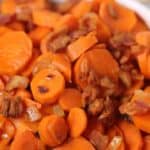 A dish of sliced cooked carrots glazed with brown sugar, mixed with small pieces of bacon, served in a white bowl.