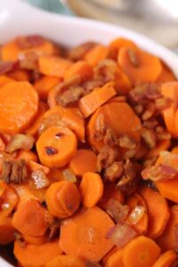 A dish of sliced cooked carrots glazed with brown sugar, mixed with small pieces of bacon, served in a white bowl.
