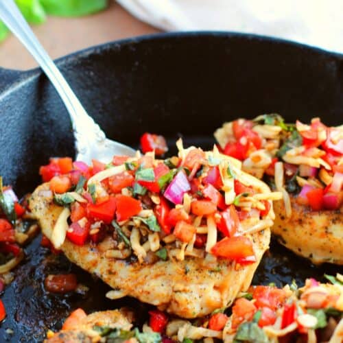 Seasoned meat in a cast iron skillet, topped with chopped tomatoes, red onion, basil, and cheese—this recipe is served with a spoon beside it. Fresh tomatoes are visible in the background.