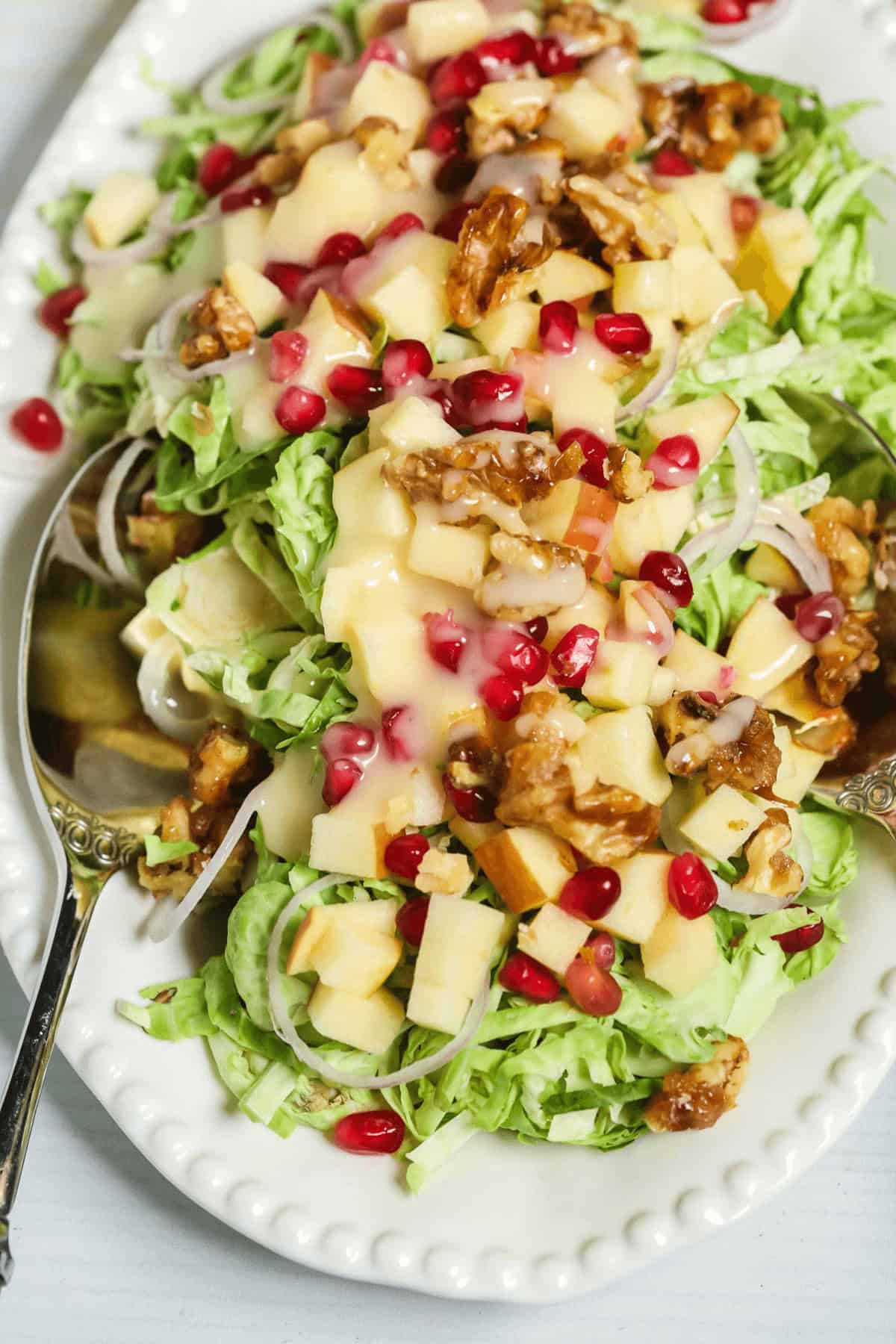 Brussels Sprouts Shaved Salad with shredded greens, diced apples, walnuts, pomegranate seeds, and creamy dressing on a white plate with a spoon.