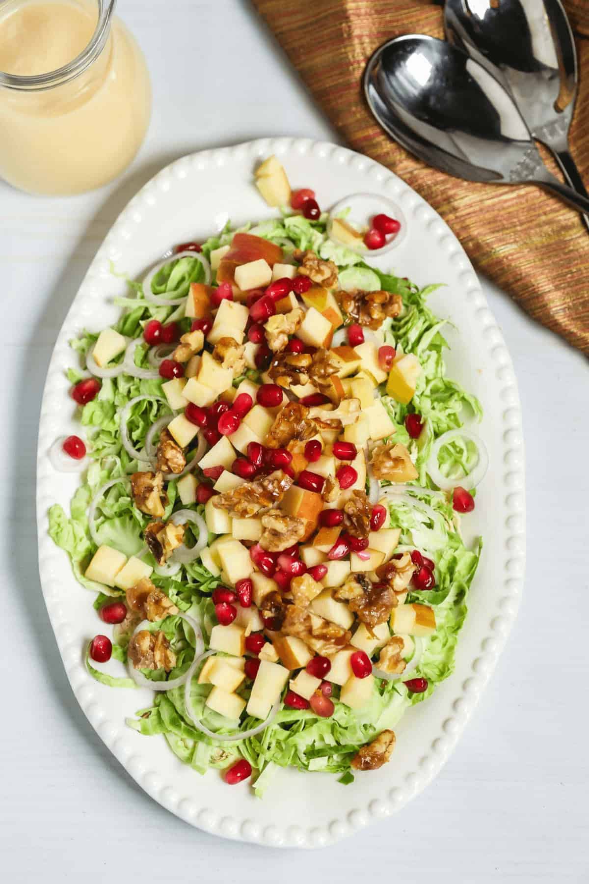 A Brussels Sprouts Shaved Salad with chopped apples, walnuts, and pomegranate seeds rests on a white oval plate. Nearby, a dressing glass and metal spoons sit gracefully on a napkin.